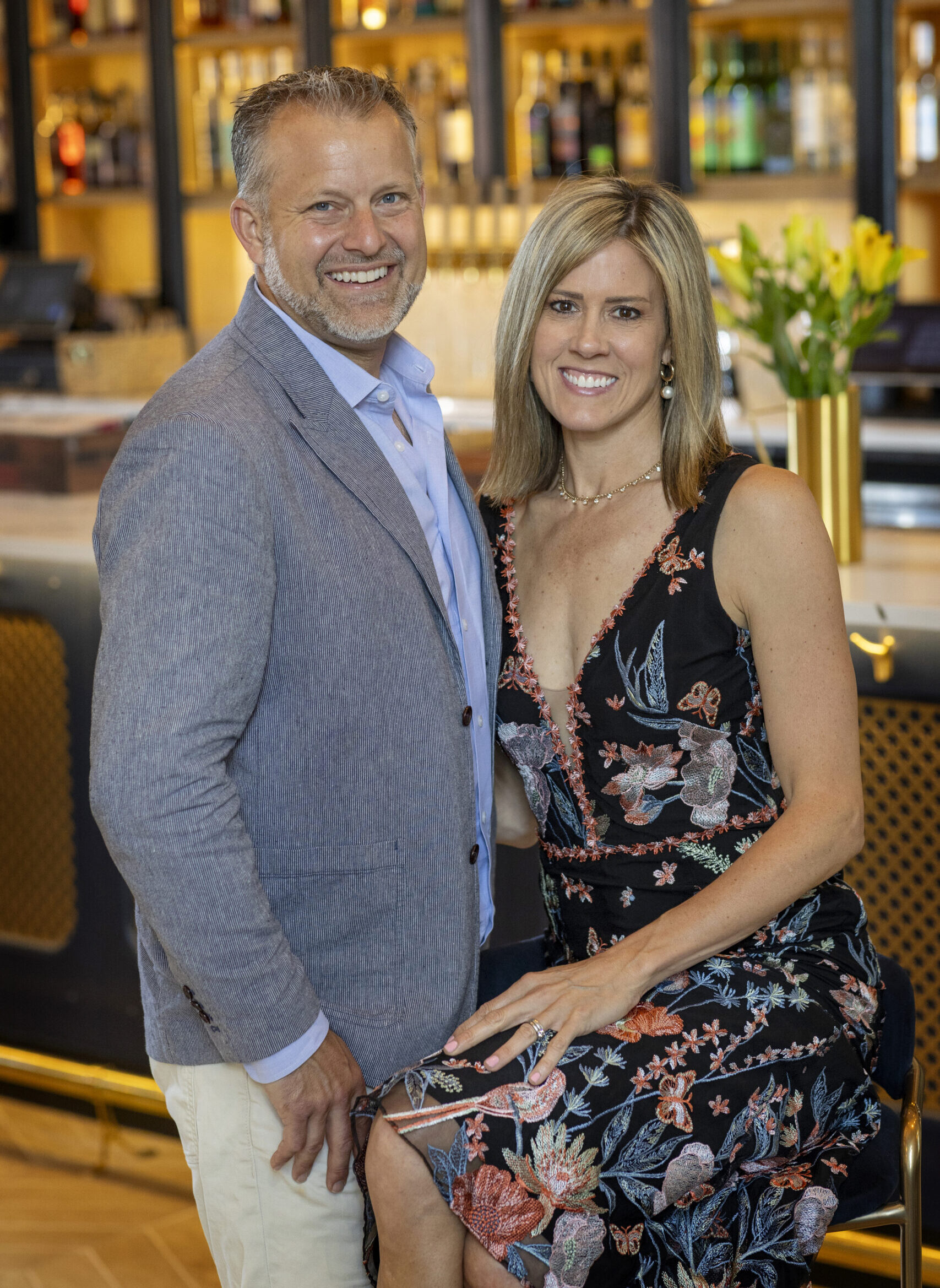 Man wearing a blazer and a woman in a dress in front of a bar smiling at the camera